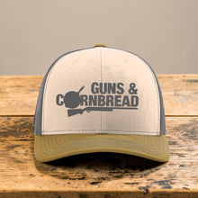 Moco Shirts Guns And Cornbread trucker hat with beige front, olive brim, and grey mesh back on a tree stump.