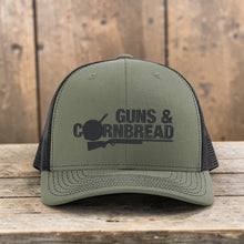 Moco Shirts Guns And Cornbread olive green trucker hat with black mesh back and graphic on a wooden stump